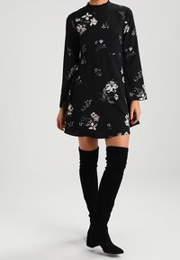 Schwarz gemustertes Mini-Kleid mit langen Ärmeln, gerafftem Ausschnitt und hellen Blumenmustern. Kombiniert mit schwarzen Overknee-Stiefeln.
