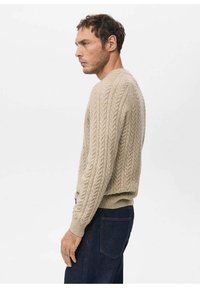 Maglione beige a trecce con polsini e orlo a costine, caratterizzato da una scollatura rotonda. Indossato con jeans in denim scuro. Vista laterale che mostra la texture e la vestibilità.