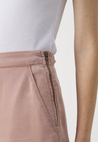 Jupe rose clair avec une texture lisse, présentant une fermeture éclair sur le côté, des poches angulaires et une taille structurée.