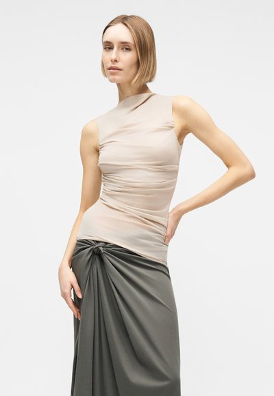 Γυναίκα με κοντά ξανθά μαλλιά, που φοράει μπεζ αμάνικο τοπ και ψηλόμεση σκουρόχρωμη γκρι φούστα με κόμπο μπροστά, ποζάροντας μπροστά σε απλό φόντο.