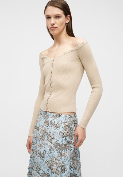 Γυναίκα που φοράει μπεζ ριμπ πλεκτή ζακέτα με ανοιχτούς ώμους και κουμπιά και μια ψηλόμεση μπλε φούστα με λουλουδάτο και ζωικό μοτίβο.