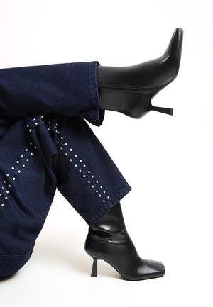 Czarne botki na obcasie sięgające kostki z ostro zakończonym noskiem i luźnym krojem. Wykonane z gładkiego materiału, w zestawieniu z ciemnymi dżinsami ozdobionymi srebrnymi ćwiekami.