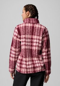 Donna con capelli intrecciati che indossa un giubbotto in pile a scacchi bordeaux e rosa, in piedi con la schiena rivolta verso uno sfondo grigio uniforme.