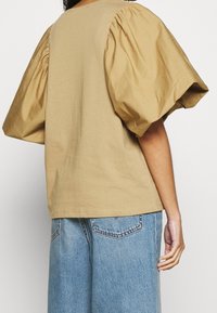 Hnědý top s nafukovacími rukávy vyrobený z měkké bavlny, s kulatým výstřihem a uvolněným střihem. Nesený s světlemodrými džíny.