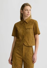 Braune, kurzärmlige Shirt mit Kragen, das eine Brusttasche und kreisförmige Ausschnitte im gesamten Stoff aufweist.