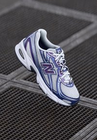 Baltas un violets New Balance sporta apavu modelis ar sieta virsu, mīkstu zoli un auklām. Sānos redzama liela logo un kontrastējoši dizaina līnijas.