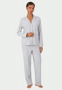 Γυναίκα που στέκεται ξυπόλητη, φορώντας σετ πιτζάμας σε ανοιχτό γκρι χρώμα με κουμπιά, μακριά μανίκια και δύο μπροστινές τσέπες.