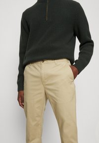 Dunkelgrüner, gerippter Strickpullover mit einem Halbzip-Halskragen, kombiniert mit beigen, leichten Baumwollhosen, die über eine Standard-Fronttasche verfügen.