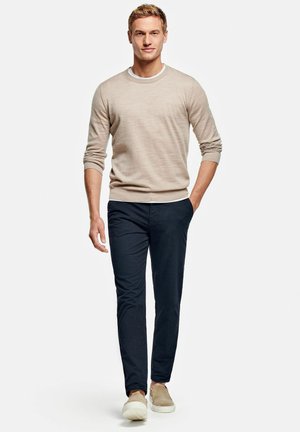 Beige Strickpullover, marineblaue Slim-Fit-Hosen und beige Slippers. Model steht mit Händen in den Taschen vor einem neutralen Hintergrund.