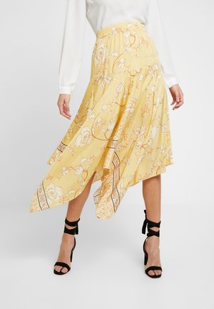 Woman wearing a yellow floral asymmetrical skirt with a white blouse and black strappy high-heeled sandals standing against a plain background.