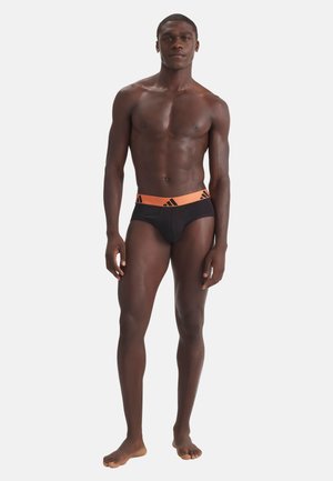 Homme athlétique à la peau foncée, debout pieds nus, portant un slip noir avec une taille élastique orange portant le logo Adidas, sur un fond blanc uni.