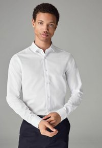 Cămașă albă cu mâneci lungi, guler cu nasturi și material texturat. Prezintă un placket frontal cu nasturi și mâneci ajustate.