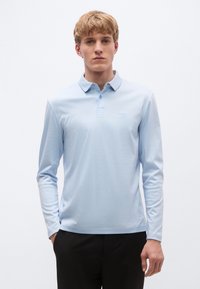 Ανοιχτό μπλε μακρυμάνικο πόλο από απαλό ύφασμα, με γιακά, τρία κουμπιά στο μπροστινό μέρος και λεπτό λογότυπο της μάρκας στο στήθος.