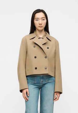 Kvinde iført en cropped beige dobbeltradet jakke over en stribet skjorte og blå jeans, stående mod en ensartet lys baggrund.