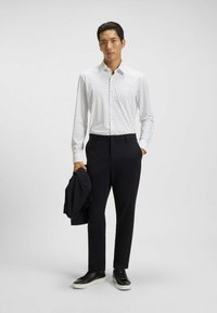Camicia bianca a maniche lunghe con piccole pois neri, abbinata a pantaloni scuri e sneakers nere. Un blazer nero è adagiato sul braccio.