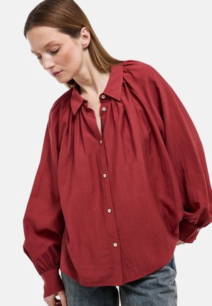 Femme portant un chemisier rouge surdimensionné à boutons avec des manches bouffantes et un jean bleu, regardant vers le bas sur un fond uni.