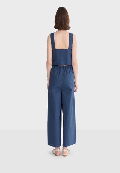Femme debout de dos, portant un crop top bleu marine sans manches et un pantalon à jambes larges, avec des sandales beiges, sur un fond uni.