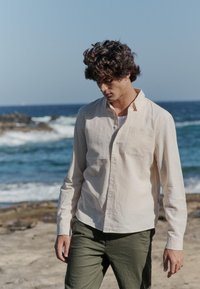 Chemise beige clair à manches longues avec col boutonné et poche poitrine, associée à un pantalon vert foncé. Arrière-plan côtier visible.