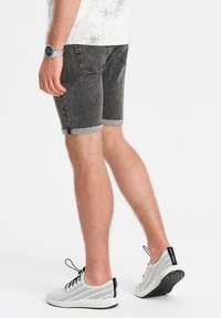 Pantalones cortos de mezclilla negros con dobladillos enrollados, que cuentan con dos bolsillos traseros. Llevados con zapatillas deportivas blancas transpirables y un reloj plateado.