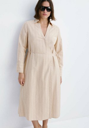 Kobieta ubrana w beżową, długorękawową, prążkowaną sukienkę midi z kołnierzykiem, okulary przeciwsłoneczne, stojąca na białym tle.