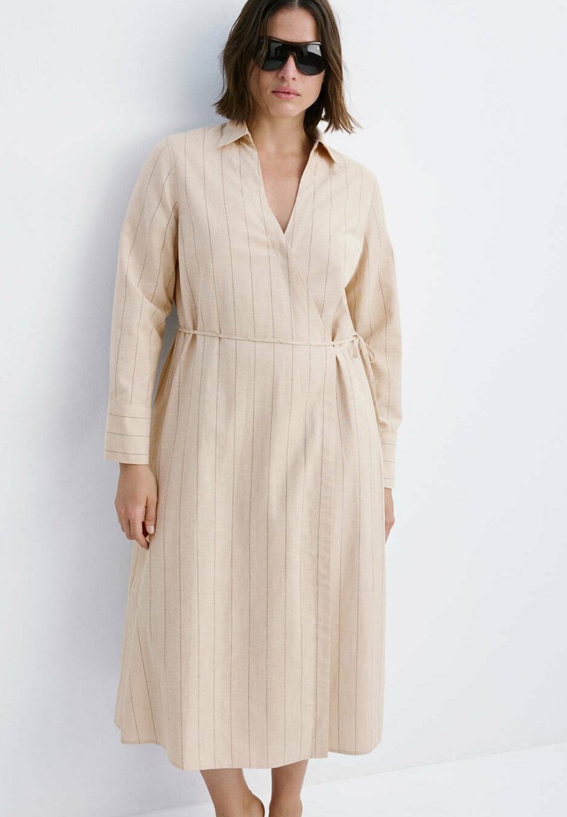 Femme portant une robe midi beige à manches longues, rayée, portefeuille avec col, lunettes de soleil, debout sur fond blanc.