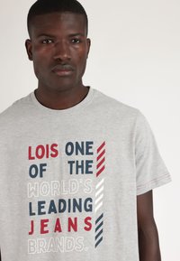 Camiseta de algodón gris con un diseño textual llamativo en rojo, azul marino y blanco, con un cuello redondo clásico y mangas cortas.