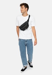 Musta nailoninen olkalaukku, jossa on vetskari, säädettävä hihna ja teksturoitu pinta, käytössä olan yli valkoisen paidan ja sinisten farkkujen päällä.