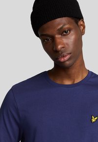 Maglietta a maniche lunghe blu navy in morbido tessuto con scollatura rotonda. Presenta un piccolo logo di un uccellino giallo sulla parte sinistra del petto.