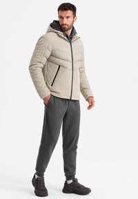 Giacca imbottita beige chiaro con design a chevron e cappuccio, abbinata a pantaloni jogger grigi e sneaker scuri. Tasche laterali con zip.