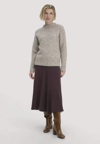 Frau in einem beigen Strickpullover, dunkellila Midirock und braunen kniehohen Stiefeln mit Absatz, stehend vor einem schlichten Hintergrund.