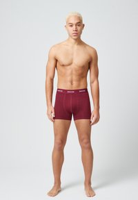 Bóxer burdeos con un ajuste ceñido, que presenta una cinturilla blanca con marca. El material parece suave y ligeramente texturizado.