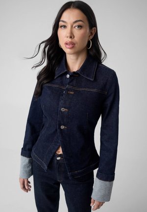 Femme aux longs cheveux foncés portant une veste en jean bleu foncé avec des poignets gris et un jean bleu foncé assorti sur un fond gris.