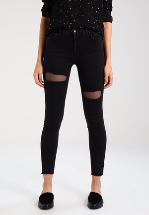 Kvinde iført sorte skinny jeans med revnede knæer, der viser netstof under, sort stjernemønstret skjorte og sorte slip-on sko med pels.