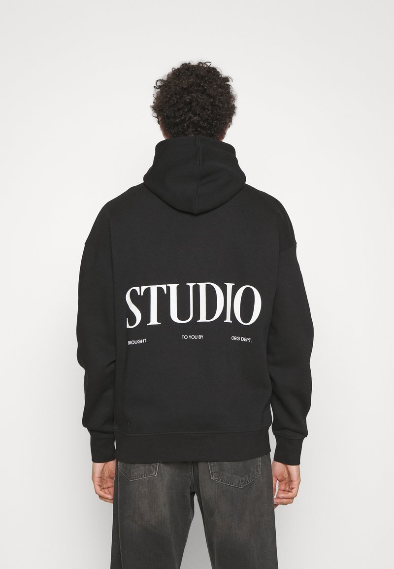 Homme aux cheveux bouclés portant un sweat à capuche noir avec un grand texte blanc « STUDIO » et un texte plus petit en dessous, debout devant un fond blanc uni.
