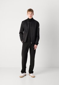 Black bomber jacket with zip closure, ribbed collar, and cuffs. Paired with black pants and white sneakers featuring tan soles.