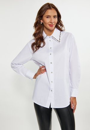 Camicia bianca con bottoni e un ampio colletto decorato con borchie argentate, maniche lunghe e orlo dritto. Abbinata a pantaloni di pelle neri.