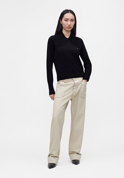 Femme debout, une main dans la poche, portant un haut noir à manches longues avec un grand col et un pantalon beige à jambes larges avec ourlets retroussés.