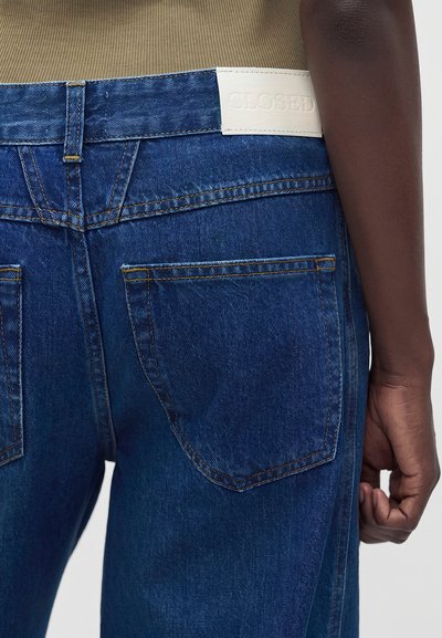 Personne à la peau foncée portant un jean en denim bleu avec des poches arrière visibles et une étiquette en cuir blanc sur la ceinture, debout avec le bras relâché.