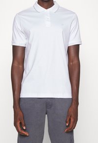 Vit pikétröja med krage och treknapparslå. Har en diskret "CK"-logotyp och märkessyntklampar på ärmarna. Mjuk, slät tyg.