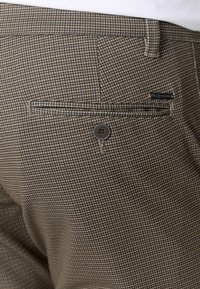 Pantaloni a quadri marroni e beige con una tasca posteriore dotata di bottone, tessuto testurizzato e una piccola etichetta del marchio sul lato destro.