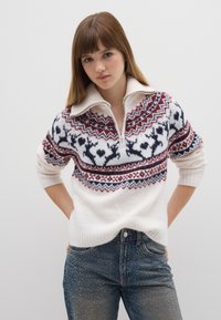 Maglione lavorato a maglia con una base bianca e motivi di renne rossi e navy, colletto alto e chiusura con zip, abbinato a jeans blu sbiaditi.
