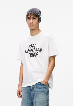 Jauns vīrietis, tērpies pelēkā beanie cepurē, baltā T-kreklā ar uzrakstu "Karl Lagerfeld Jeans" un zilos džinsos, stāvot ar vienu roku kabatā.