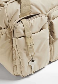 Borsa imbottita beige con più tasche con zip, tessuto testurizzato, tracolla regolabile e una chiave attaccata a un anello metallico.