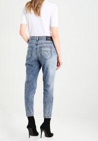 Nainen, jolla on päällään vaaleansiniset korkeavyötäröiset farkut, valkoinen vartalonmyötäinen lyhythihainen paita ja mustat korkean kantapään nilkkakengät, seisoo selkä kameralle.