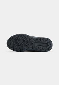 Semelle de chaussure noire avec des crampons profonds et des motifs géométriques conçus pour l’adhérence, présentée sur un fond gris clair uni.