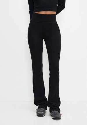 Woman wearing black high-waist flare pants and grey sneakers, standing against a white background with hands behind back.