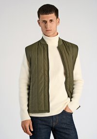 Olivgrüne, gesteppte Weste über einem cremefarbenen Rollkragenpullover, mit durchgehendem Reißverschluss und gerippten Bündchen, kombiniert mit dunkelblauen Jeans.