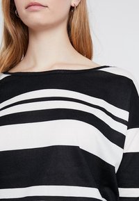 Gestreiftes Oberteil in Schwarz und Weiß mit weitem Ausschnitt, aus weichem Baumwollmaterial und mit kurzen Ärmeln. Die Streifen variieren in der Breite. Einfaches Design.