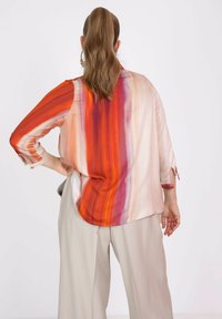 Zijden blouse met een verloop van oranje, rood en paarse strepen, lange mouwen en een ronde zoom, gecombineerd met lichtbeige broek.
