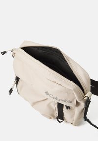 Beige Columbia skuldertaske med en lynlås i toppen, sort indvendig foring, udvendige lynlåslommer og justerbar sort rem.